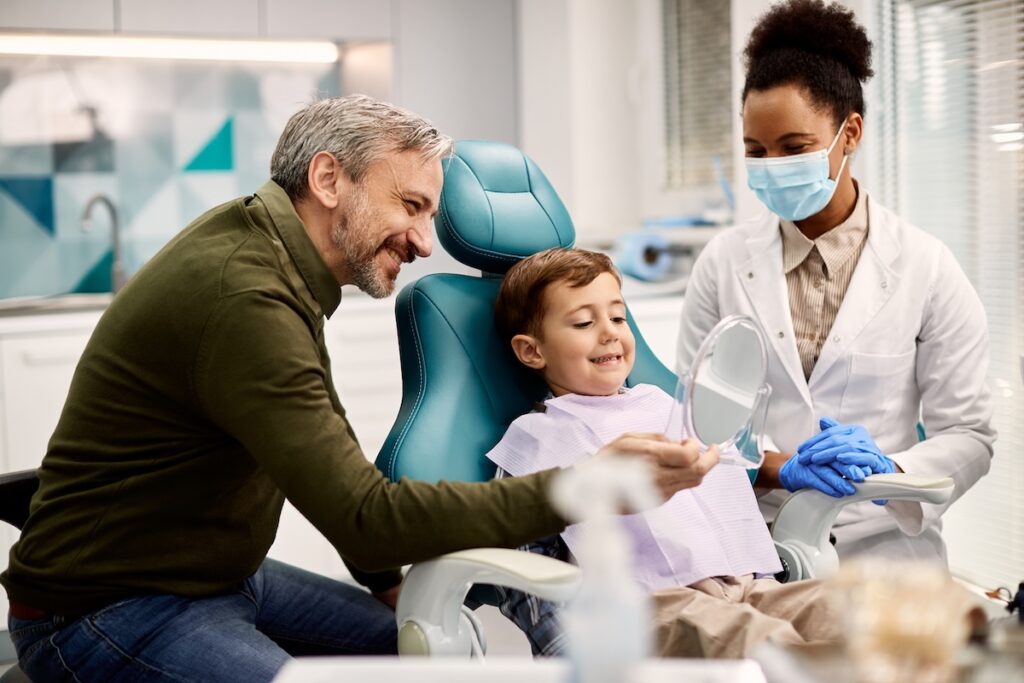 Happy father holding a mirror while son is looking at his teeth after dental appointment at dentist's office.