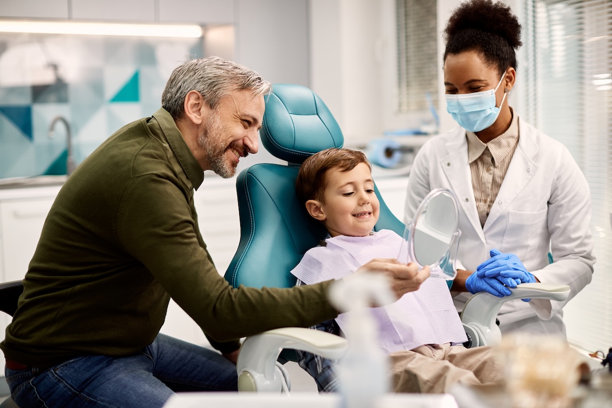 Happy father holding a mirror while son is looking at his teeth after dental appointment at dentist's office.